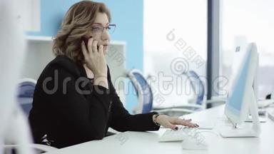 年轻的休闲女士坐在办公桌前，用笔记本电脑，接电话，打电话，微笑着在手机上说话